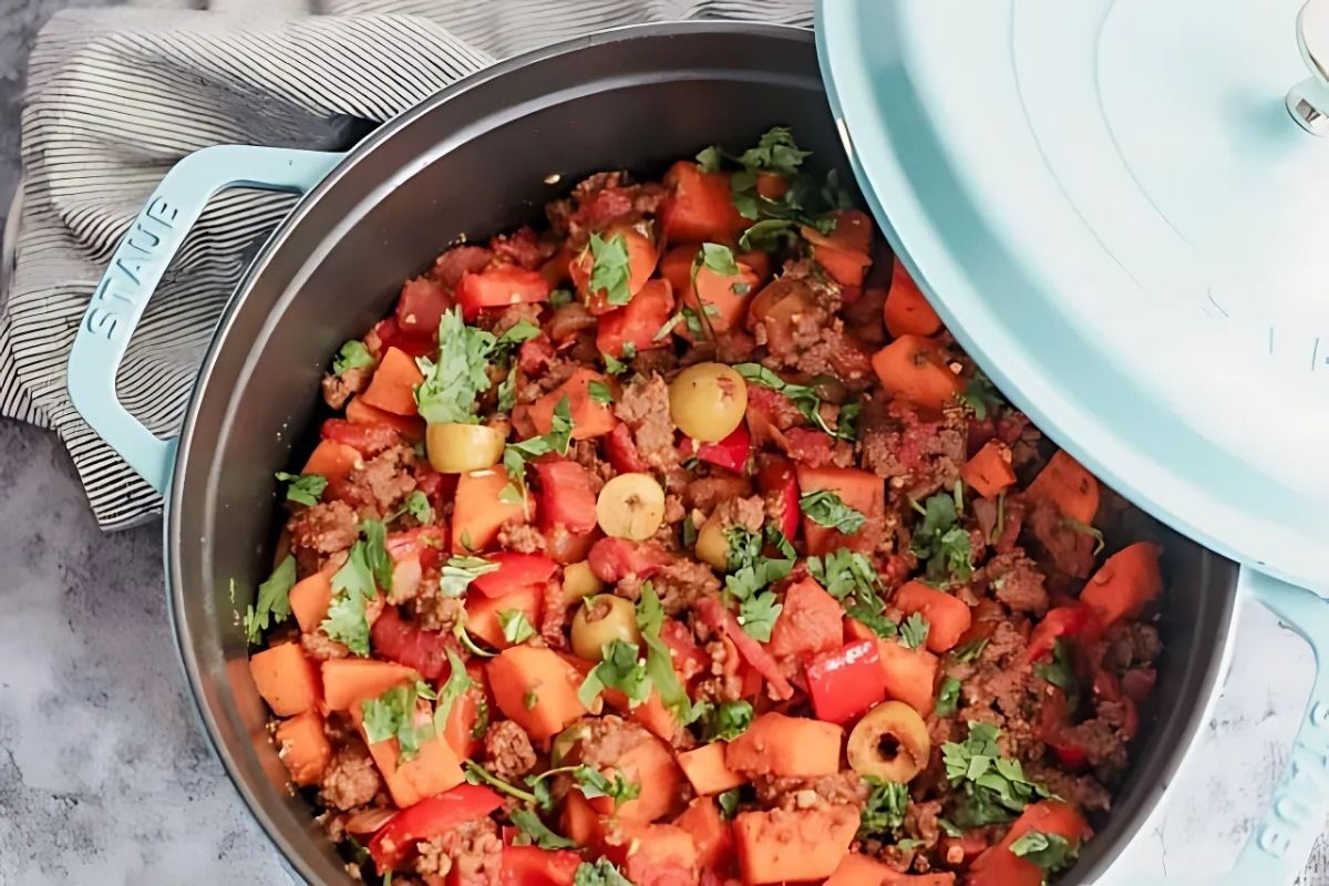 Overhead view of a light‑blue cast‑iron skillet brimming with Costa Rican sweet‑potato picadillo—crumbled grass‑fed beef, orange sweet‑potato cubes, red bell peppers, tomatoes, and green olives—garnished with fresh cilantro.