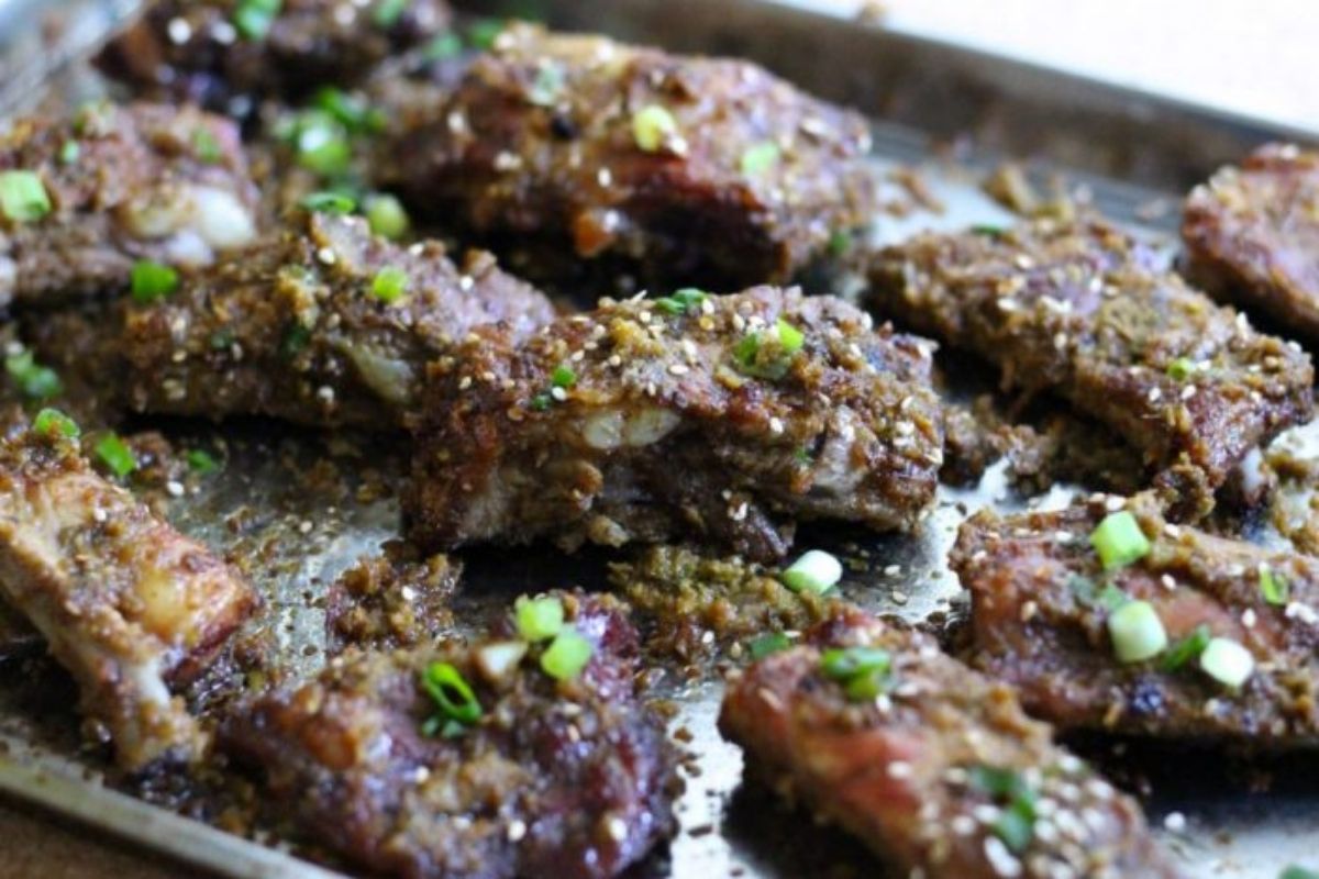 Close-up of juicy, oven-baked Paleo Korean pork ribs on a metal baking sheet, coated in a caramelized, savory sauce and sprinkled with sesame seeds and chopped green onions.