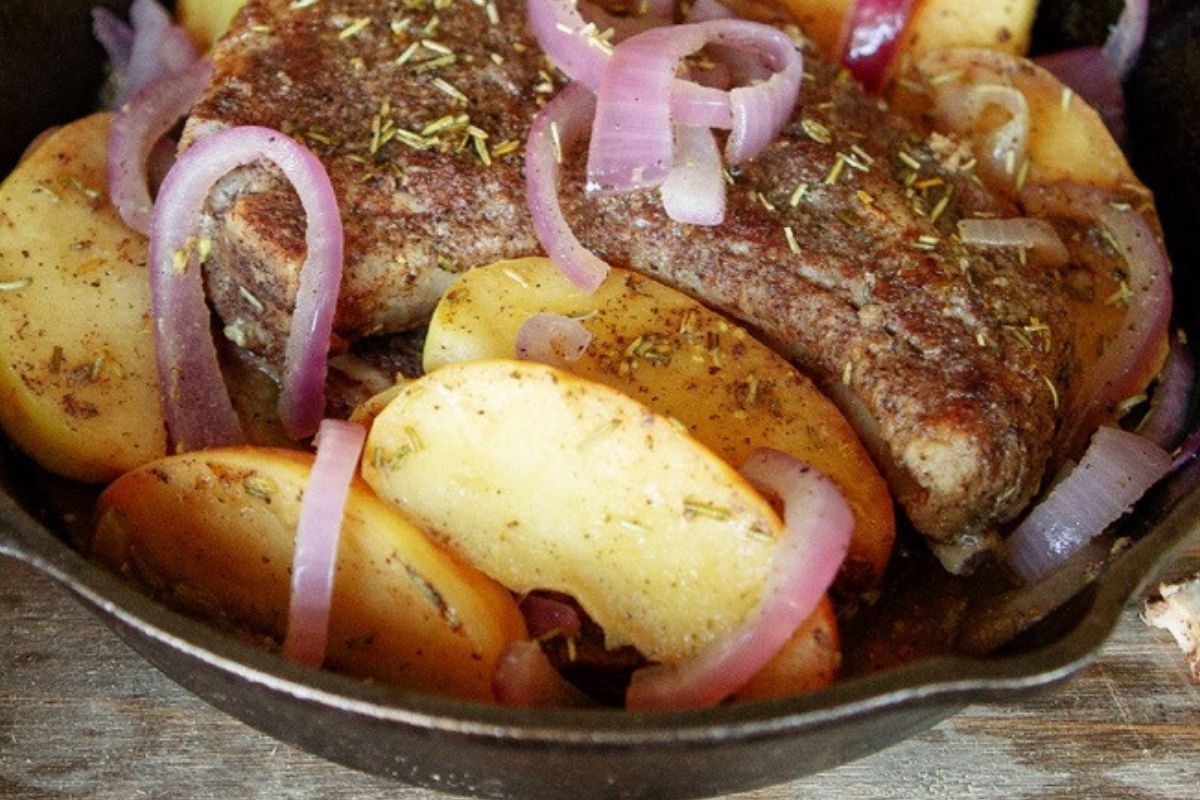 Close-up of seared boneless pork chops in a cast iron skillet, surrounded by sautéed red onions and golden apple slices, seasoned with herbs.