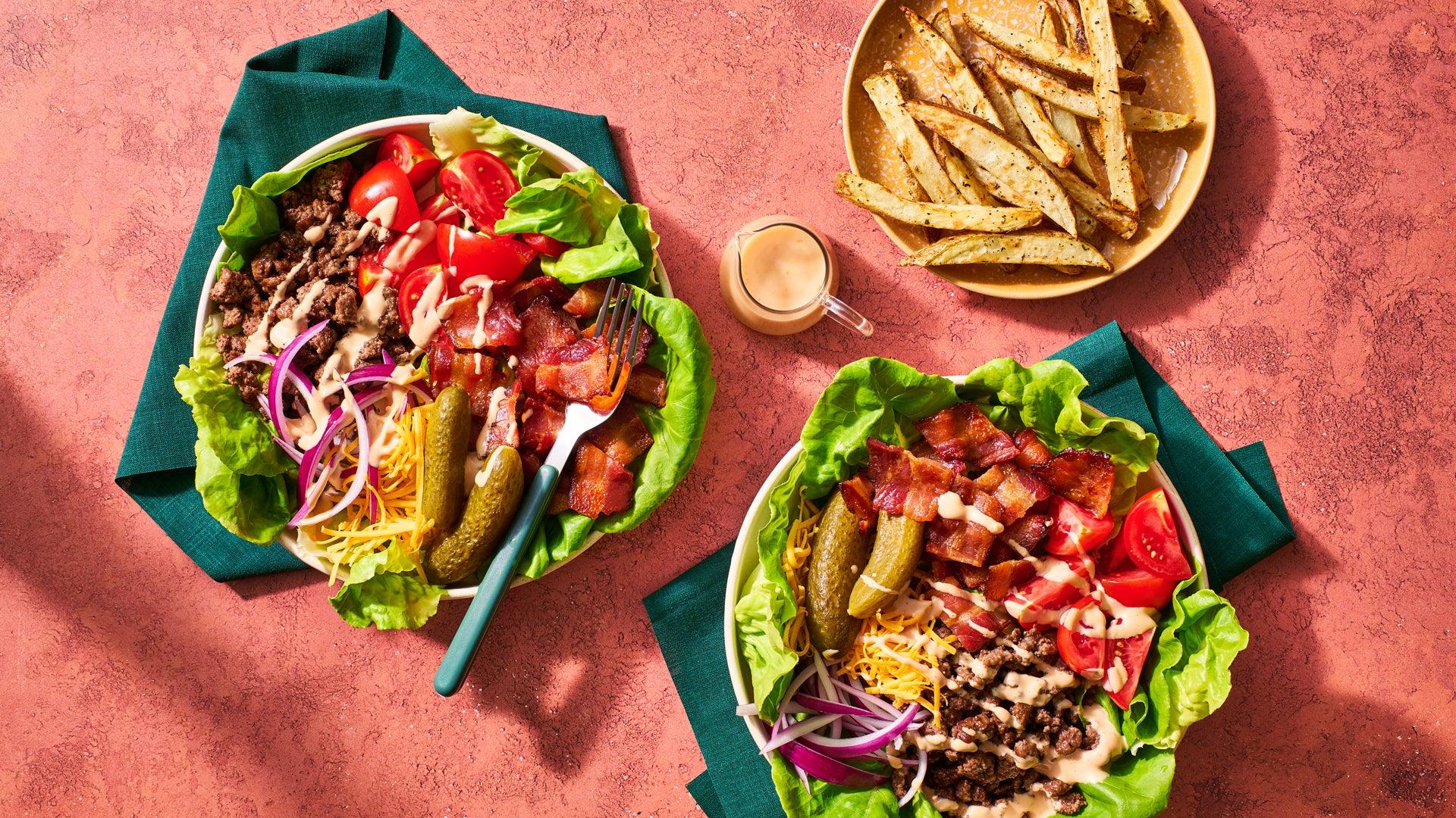 Two bowls of loaded beef and cheese salad with lettuce, tomatoes, onions, pickles, bacon bits, and dressing, served on dark green napkins on a textured pink surface. A plate of potato wedges and a small bottle of dressing are also shown.