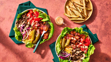 Two bowls of loaded beef and cheese salad with lettuce, tomatoes, onions, pickles, bacon bits, and dressing, served on dark green napkins on a textured pink surface. A plate of potato wedges and a small bottle of dressing are also shown.