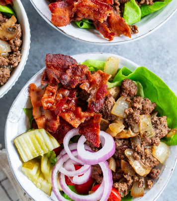 Three bowls of a gourmet breakfast salad featuring crispy bacon, cooked ground beef with caramelized onions, green lettuce, pickles, and sliced red onion on a gray background.