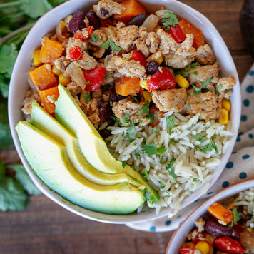 A closeup of a bowl of cooked ground pork mixed with bell peppers. Beside it is seasoned rice and fresh avocado slices.