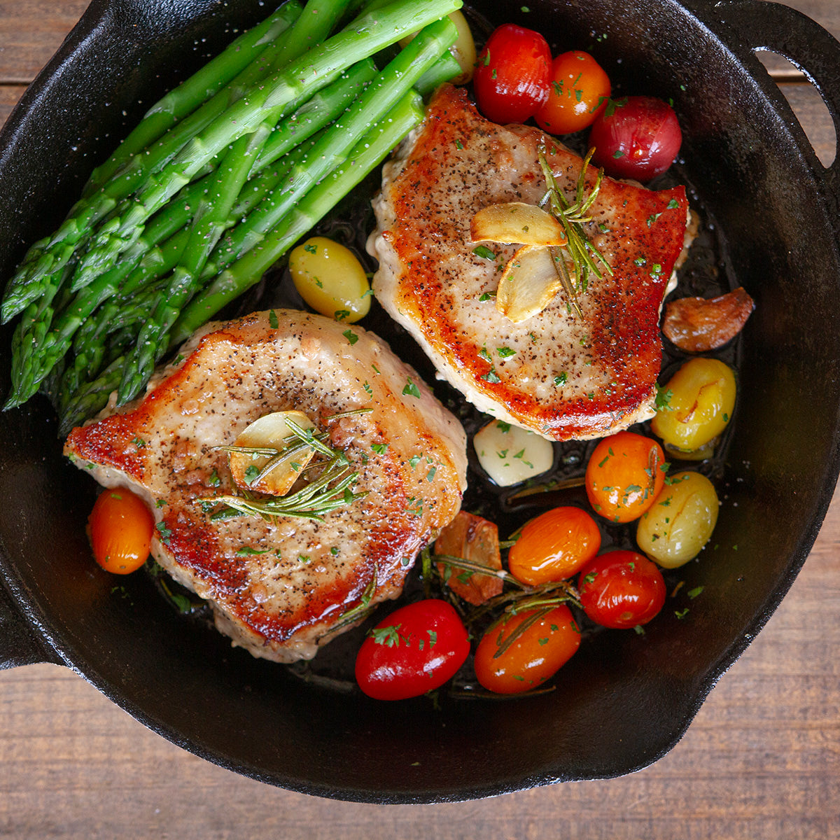 Two grilled thick cut pork chops sit in a cast iron pan surrounded by cooked asparagus and red, yellow, and orange cherry tomatoes.