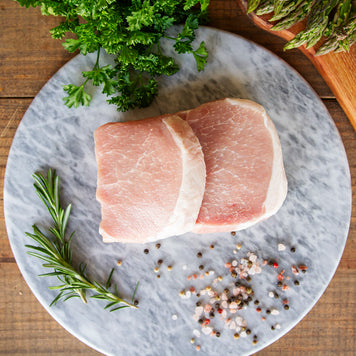 Two thick cut boneless pork chops sit on a marble cutting board with fresh herbs and peppercorns around the edges.
